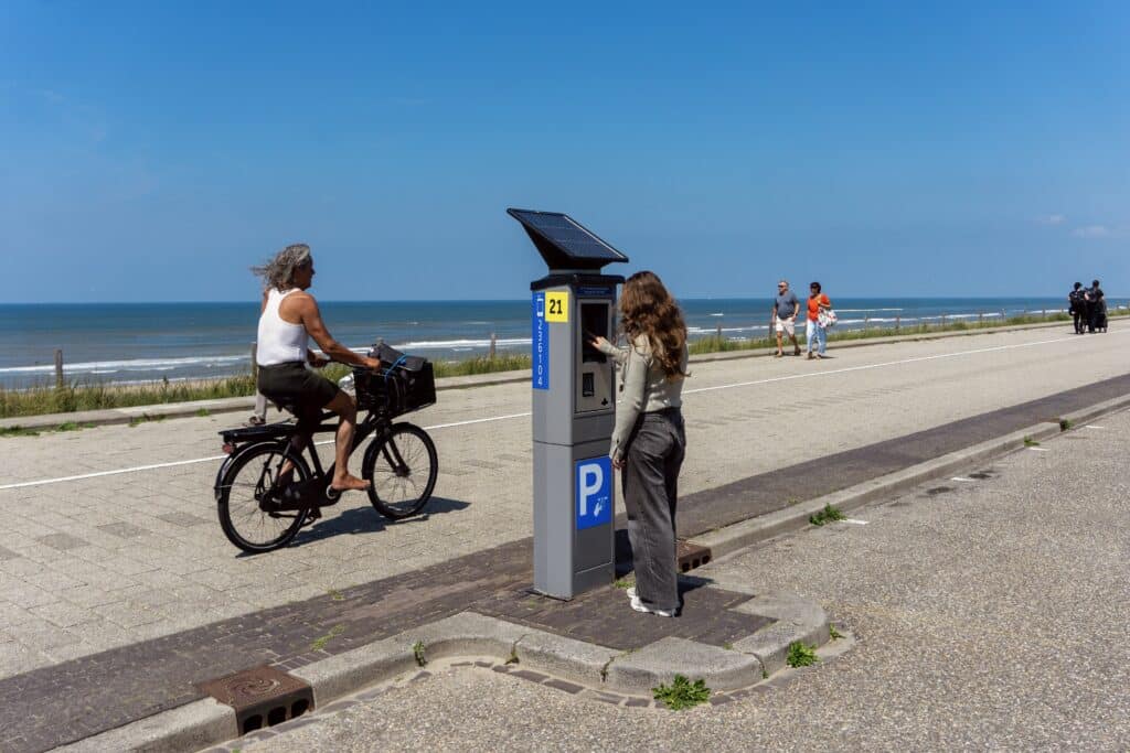 Person nutzt einen Parkautomaten auf dem Boulevard von Zandvoort, während ein Radfahrer mit Blick auf Strand und Meer vorbeifährt.